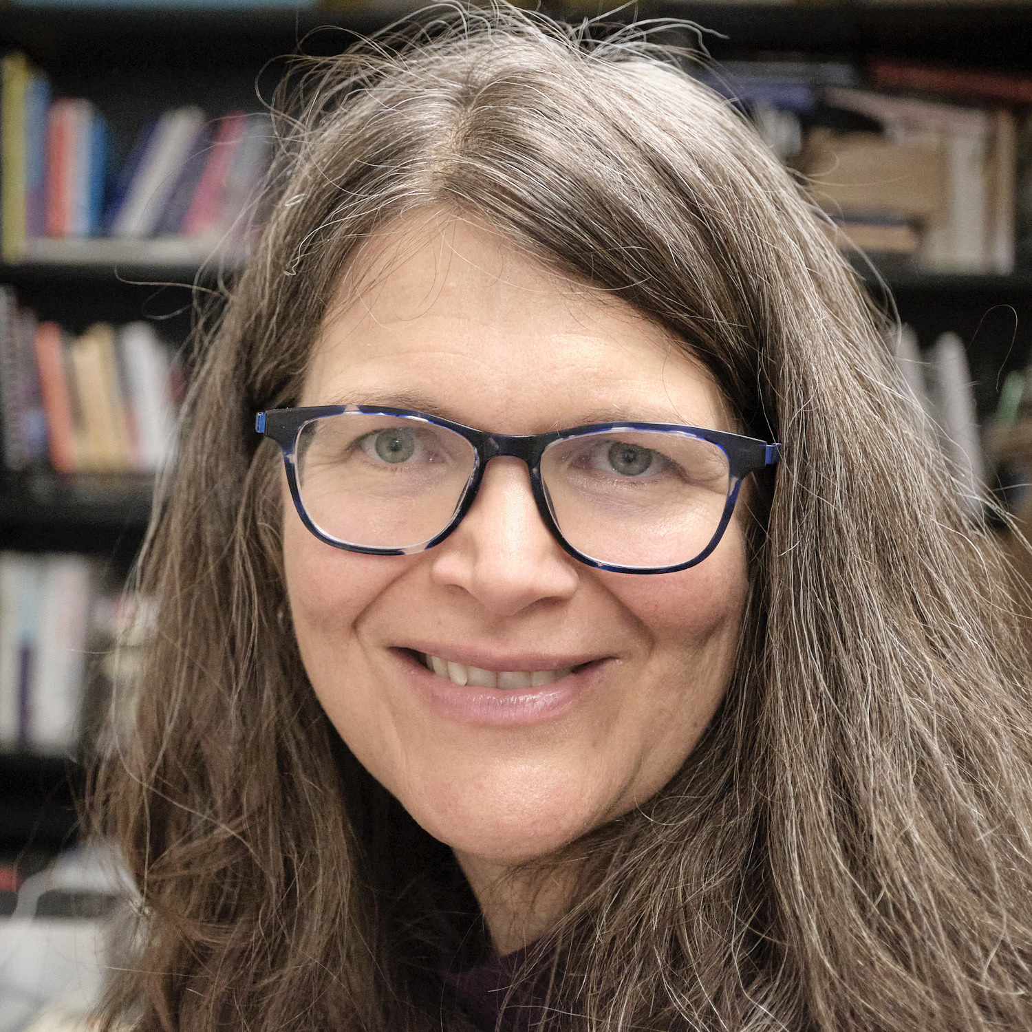 Smiling woman with large blue glasses and long brown hair.